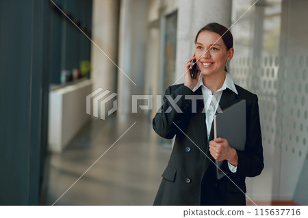 A professional woman in office is talking on phone while holding a folder and smiling A professional woman in office is talking on phone while holding a folder and smiling 115637716