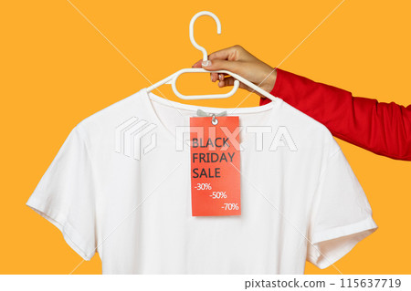 A hand is shown holding a white plastic hanger, which supports a plain white t-shirt featuring a vibrant red sale tag advertising Black Friday discounts, cropped A hand is shown holding a white plastic hanger, which supports a plain white t-shirt featuring a vibrant red sale tag advertising Black Friday discounts, cropped 115637719