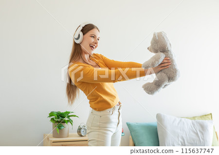 A girl is seen holding a teddy bear while listening to music through headphones. She appears relaxed and focused, enjoying the music playing in her ears. 115637747