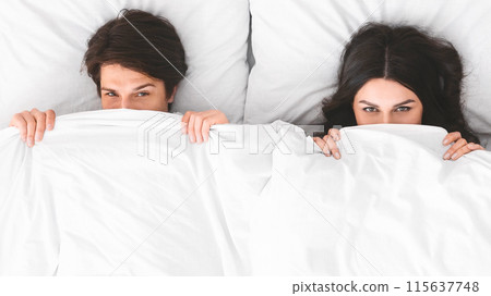 A man and a woman are lying in bed, both partially hidden under a white blanket. They are both looking at the camera with a playful, mischievous expression. 115637748