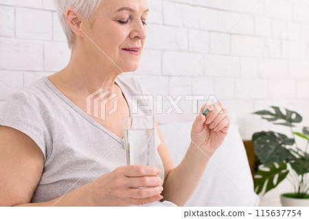 Senior woman with short grey hair is gently holding a pill in one hand and a clear glass of water in the other. She appears calm and focused, preparing to take her medication after wake up 115637754