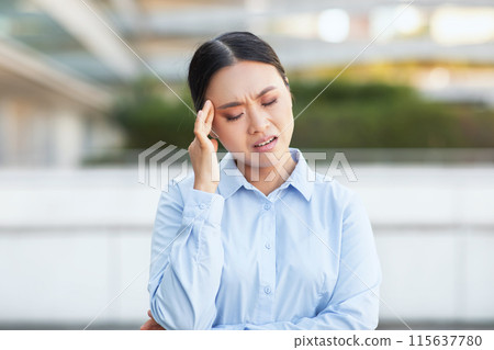 Asian woman is shown holding her head in a gesture of pain or distress, her facial expression suggesting discomfort. She appears to be experiencing a headache or migraine Asian woman is shown holding her head in a gesture of pain or distress, her facial expression suggesting discomfort. She appears to be experiencing a headache or migraine 115637780