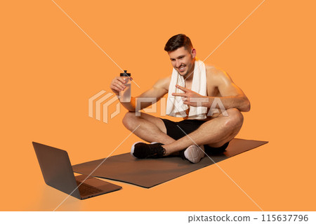 A young man is sitting cross-legged on a yoga mat, engaging in a virtual workout session. He wears a towel around his neck and holds a water bottle, smiling and gesturing towards his open laptop. 115637796