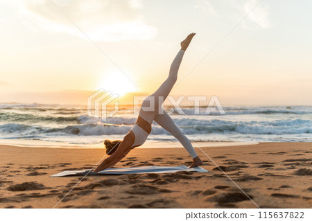 Young woman in sportswear doing yoga exercise with raised leg outdoors, having workout on sea beach at sunset, side view, full length. Lady stretching outside 115637822