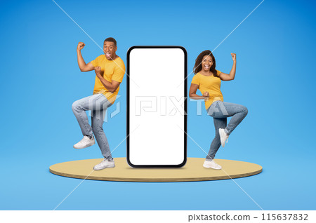 Emotional happy young african american couple in casual gesturing and exclaiming, standing on platform with huge phone with white blank screen over blue background, mockup, online gambling, bet 115637832