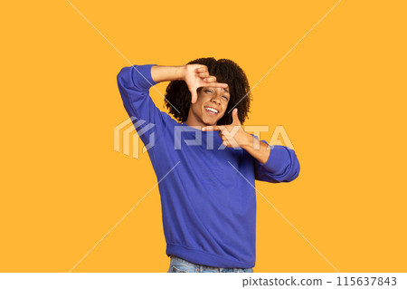 A young man with curly dark hair is wearing a blue sweatshirt and is standing against a yellow background. He is looking directly at the camera and smiling while making a framing gesture A young man with curly dark hair is wearing a blue sweatshirt and is standing against a yellow background. He is looking directly at the camera and smiling while making a framing gesture 115637843