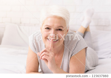 A cheerful senior woman with short white hair lies on her stomach on a bed, her eyes beaming with joy. She places her finger on her lips, suggesting a playful secret or a quiet moment A cheerful senior woman with short white hair lies on her stomach on a bed, her eyes beaming with joy. She places her finger on her lips, suggesting a playful secret or a quiet moment 115637860