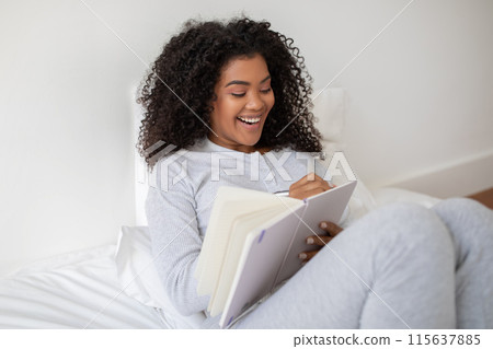 A cheerful young Hispanic woman is seen comfortably sitting in bed with her legs crossed, deeply engaged and smiling as she write in notebook 115637885