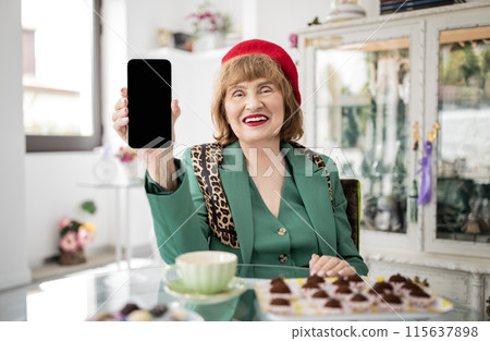 Cheerful Senior Lady Showing Smartphone Blank Screen Advertising Application For Online Fun Sitting At Cafe Interior, Wearing Bright Stylish Clothes, Recommending Mobile Offer. Mockup Cheerful Senior Lady Showing Smartphone Blank Screen Advertising Application For Online Fun Sitting At Cafe Interior, Wearing Bright Stylish Clothes, Recommending Mobile Offer. Mockup 115637898