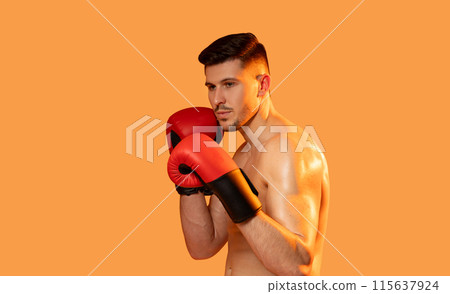 A man wearing boxing gloves strikes a pose against a vibrant orange background. He exudes confidence and strength as he showcases his boxing stance. A man wearing boxing gloves strikes a pose against a vibrant orange background. He exudes confidence and strength as he showcases his boxing stance. 115637924