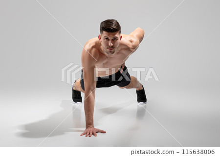 A fit young man is captured performing a plank exercise in a minimalist studio bathed in sunlight. He appears focused and determined, showcasing his athletic build and dedication to fitness. 115638006