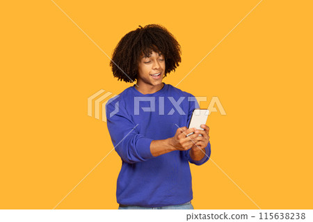A young man with curly hair is wearing a blue shirt and smiling as he holds a phone in his hands. The backdrop is a vivid orange color which enhances the cheerful atmosphere of the scene. A young man with curly hair is wearing a blue shirt and smiling as he holds a phone in his hands. The backdrop is a vivid orange color which enhances the cheerful atmosphere of the scene. 115638238