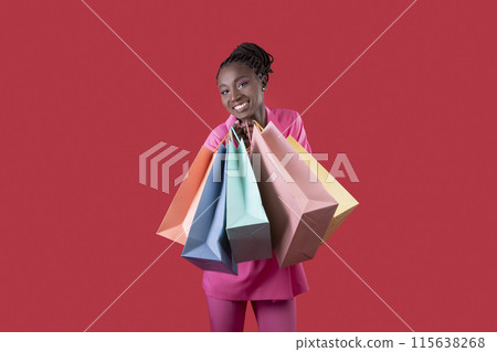 Beautiful Smiling African American Woman With Shopping Bags In Hands Posing Over Red Studio Background, Happy Black Shopaholic Lady In Pink Suit Looking At Camera, Enjoying Seasonal Sales, Copy Space 115638268