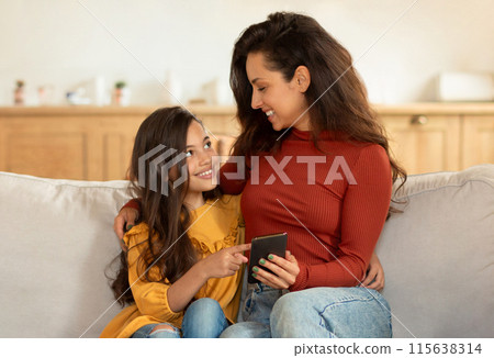 Happy Middle Eastern Mom And Little Daughter Using Smartphone, Kid Girl Pointing Finger At Gadget, Showing Her Favorite Mobile Game Application, Hugging Sitting On Couch At Home Interior 115638314