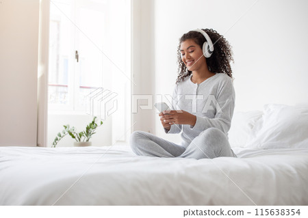 A young Hispanic woman sits cross-legged on her bed, wearing comfortable white pajamas and wireless headphones, engaged with her smartphone, possibly choosing a song or responding to a message A young Hispanic woman sits cross-legged on her bed, wearing comfortable white pajamas and wireless headphones, engaged with her smartphone, possibly choosing a song or responding to a message 115638354