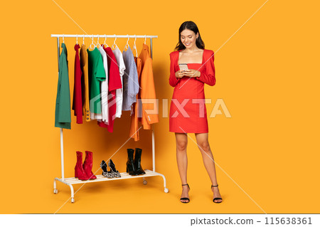 A young woman stands with a content smile on her face as she browses her smartphone. A rack of colorful clothing is to her left, with various garments neatly hung and pairs of boots placed beneath 115638361