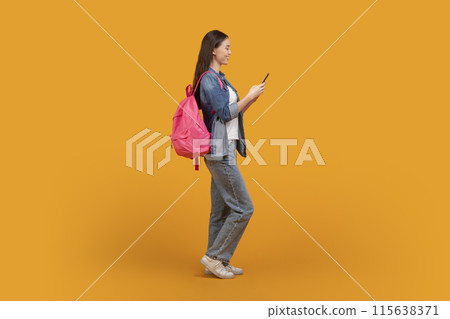 A young woman with long dark hair is wearing a blue jean jacket and jeans. She is carrying a pink backpack and using a smartphone. She is standing on a yellow background. 115638371