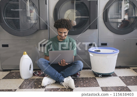 Easy Laundry. Happy Hispanic Student Guy Using Laptop While Washing Clothes At Laundromat, Studying Online Sitting Near Industrial Washer Machines, Detergent Bottle And Basket With Clothing Indoor 115638403