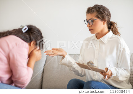 A therapist, wearing a white shirt and glasses, sits on a couch and gestures with her right hand while speaking to a patient. The patient, with their head down, appears to be crying or distressed A therapist, wearing a white shirt and glasses, sits on a couch and gestures with her right hand while speaking to a patient. The patient, with their head down, appears to be crying or distressed 115638454