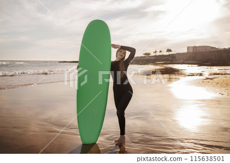 Beautiful young female surfer posing with her surfboard at the beach on sunset, portrait of happy millennial woman in wetsuit surfing at ocean shore, enjoying water sports, full length shot Beautiful young female surfer posing with her surfboard at the beach on sunset, portrait of happy millennial woman in wetsuit surfing at ocean shore, enjoying water sports, full length shot 115638501
