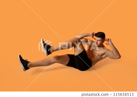 A fit man in black shorts and sneakers is performing a bicycle crunch exercise on an orange studio background. He is focused and demonstrating proper form. 115638513
