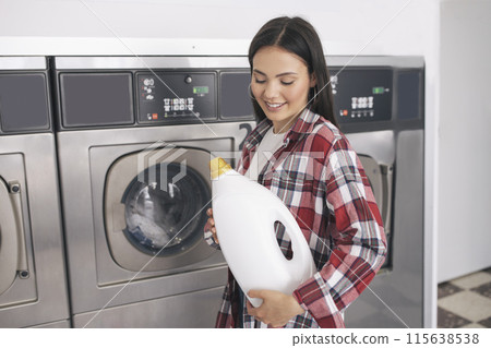 Laundromat. Young Woman Smiling Holding Detergent Bottle, Cleaning And Washing Clothes At Public Laundrette Service, Standing At Row Of Industrial Washer Machines Indoors 115638538