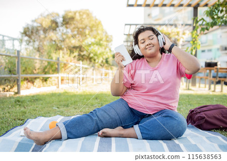 Happy Overweight Latin Woman Listening To Music Online With Headphones And Smartphone, Sitting On Blanket Enjoying Picnic In Park Outside. Lady Closing Eyes, Listens Favorite Playlist On Summer Day Happy Overweight Latin Woman Listening To Music Online With Headphones And Smartphone, Sitting On Blanket Enjoying Picnic In Park Outside. Lady Closing Eyes, Listens Favorite Playlist On Summer Day 115638563