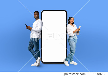 On a deep blue background, a multiracial couple, both smiling, stands adjacent to an enormous smartphone with a blank display, ready for showcasing apps or designs. On a deep blue background, a multiracial couple, both smiling, stands adjacent to an enormous smartphone with a blank display, ready for showcasing apps or designs. 115638612