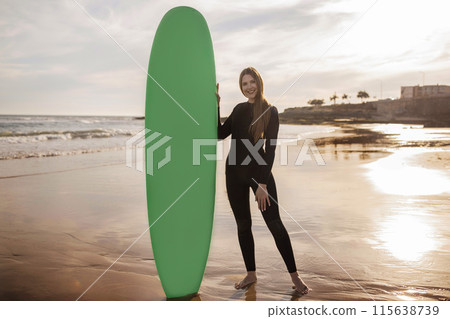 Professional Surfer Lady Standing On The Beach And Holding Surf Board, Beautiful Young Woman In Wetsuit Surfing On Sunset Time, Waiting For High Waves, Looking At Camera And Smiling, Copy Space 115638739