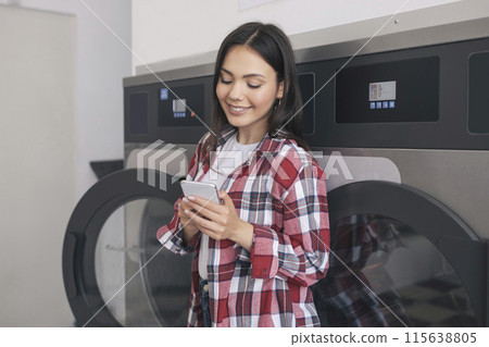 Happy Student Woman Doing Laundry And Texting On Cellphone, Booking Laundromat In App While Washing Her Clothes At Public Laundrette Service Indoors, Standing Near Row Of Washer Machines Happy Student Woman Doing Laundry And Texting On Cellphone, Booking Laundromat In App While Washing Her Clothes At Public Laundrette Service Indoors, Standing Near Row Of Washer Machines 115638805