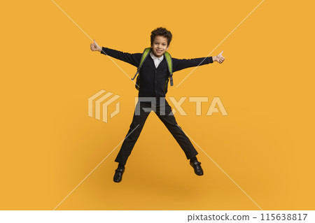 Black school boy, wearing a black uniform and a green backpack, jumps in the air with a big smile. He has his arms outstretched and is giving a thumbs up with both hands. 115638817