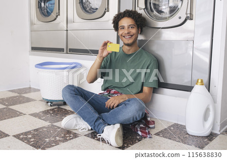 Young Latin Man Showing Credit Card Sitting With Detergent Bottle At Laundry Room Indoor, Smiling At Camera Posing Near Row Of Industrial Washing Machines. Great Public Conveniences Offer 115638830