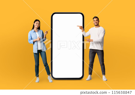 The scene features a young multiethnic couple, both grinning, positioned next to a life-sized smartphone mockup on a seamless yellow background. The scene features a young multiethnic couple, both grinning, positioned next to a life-sized smartphone mockup on a seamless yellow background. 115638847