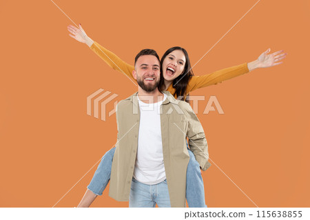 A cheerful young man in casual attire gives a piggyback ride to a happy young woman, both expressing joy with bright smiles and raised arms against an orange background. 115638855