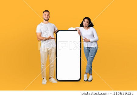 A man and a woman are standing together, holding a blank phone in their hands. They are posing against a bright yellow background, looking directly at the camera. 115639123