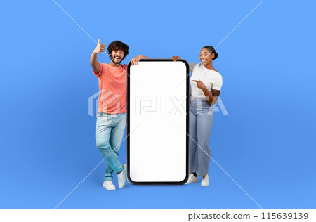 A cheerful man and woman stand in a studio against a vibrant blue background. They are holding a large blank smartphone banner between them. 115639139