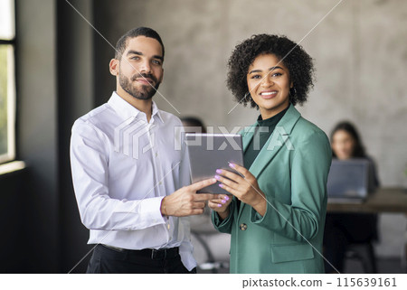 Two Diverse Business Specialists Using Digital Tablet Together, Standing Smiling At Camera In Modern Office Interior. Arabic Businessman And Black Businesswoman Websurfing At Workplace Two Diverse Business Specialists Using Digital Tablet Together, Standing Smiling At Camera In Modern Office Interior. Arabic Businessman And Black Businesswoman Websurfing At Workplace 115639161