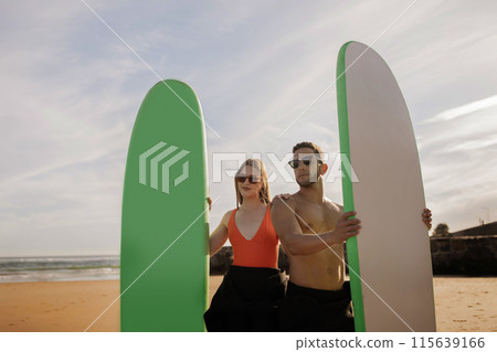 Young Beautiful Couple With Surf Boards In Hands Posing On The Beach In Summer, Smiling Men And Women Wearing Stylish Sunglasses Holding Surfboards And Looking Away, Surfers Friends Ready For Surfing Young Beautiful Couple With Surf Boards In Hands Posing On The Beach In Summer, Smiling Men And Women Wearing Stylish Sunglasses Holding Surfboards And Looking Away, Surfers Friends Ready For Surfing 115639166