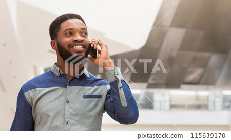 A black man is actively engaged in a phone conversation while standing outdoors. He appears focused and attentive as he communicates via his cell phone, closeup, copy space A black man is actively engaged in a phone conversation while standing outdoors. He appears focused and attentive as he communicates via his cell phone, closeup, copy space 115639170