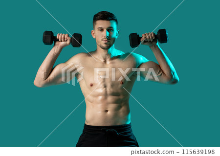 A man stands in front of a vibrant green background, gripping two dumbbells in his hands. He appears focused and determined, showcasing strength and fitness. 115639198