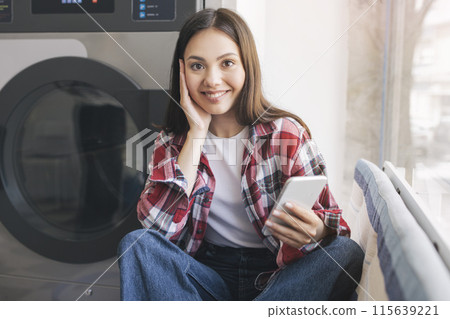 Laundry. Happy European Woman With Cellphone Sitting Near Washing Machine Indoor, Smiling At Camera. Young Lady Booking Laundromat Online. Modern Technology And Laundrette Chores Concept 115639221