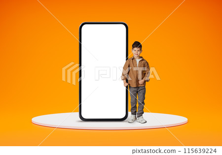 Educational kids mobile app. Happy positive cute little boy standing by big smartphone with black white screen and smiling, ready for school, posing over orange studio background, copy space Educational kids mobile app. Happy positive cute little boy standing by big smartphone with black white screen and smiling, ready for school, posing over orange studio background, copy space 115639224