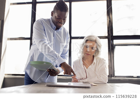 Cheerful Black Businessman And Mature Colleague Lady Working Together On Business Project, Man Pointing Finger At Papers On Table In Modern Office Interior. Teamwork And Coworkers Cooperation 115639228