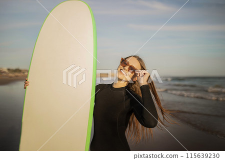 Portrait Of Beautiful Young Surfer Lady Holding Big Blank Surfboard, Smiling Millennial Woman Wearing Stylish Sunglasses And Wetsuit Standing On The Beach At Sunset, Enjoying Surfing, Copy Space 115639230
