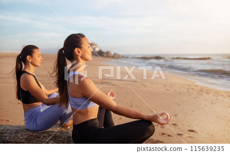 Calm cheerful young caucasian women enjoy morning workout, sunrise, spare time, vitality, breathing exercises and practice yoga in lotus position on sea beach. Meditation, hobby outdoor 115639235