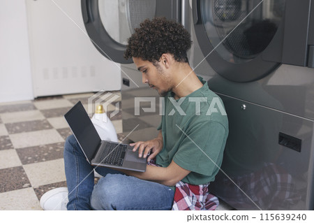 Great Laundry Service. Middle Eastern Student Guy Learning Online Via Laptop While Washing Clothes At Public Laundrette, Sitting Indoor With Computer And Surfing Web, Wearing Casual. Side View 115639240