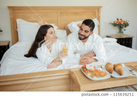Hotel Room Service. Relaxed Couple Enjoying Romantic Breakfast In Bed, Drinking Fresh Orange Juice Lying Wearing Bathrobes In Bedroom Interior, Near Tray With Delicious Food. Honeymoon Vacation 115639251
