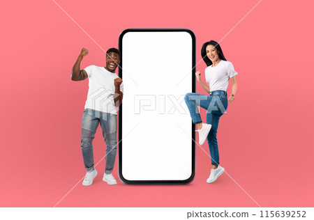 A smiling multiethnic young man and woman stand confidently beside a large smartphone with a blank screen, isolated on a vivid blue background. A smiling multiethnic young man and woman stand confidently beside a large smartphone with a blank screen, isolated on a vivid blue background. 115639252