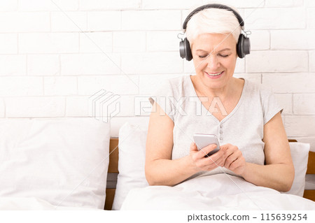 Senior woman listening to music on his smartphone at home in bed, using modern wireless headphones, panorama, copy space Senior woman listening to music on his smartphone at home in bed, using modern wireless headphones, panorama, copy space 115639254