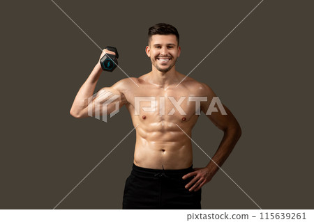A man is standing and posing while holding a dumbbell in his hand. He is showcasing his muscles and strength, with a focused expression on his face. A man is standing and posing while holding a dumbbell in his hand. He is showcasing his muscles and strength, with a focused expression on his face. 115639261
